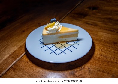 Slice Of Cheesecake With Lemon Slice And Blueberry, Topped With Honey On White Plate. Dark Tone Background.