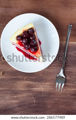 Slice of cheesecake topped with cherry compote on plate on brown table with dessert fork, top view