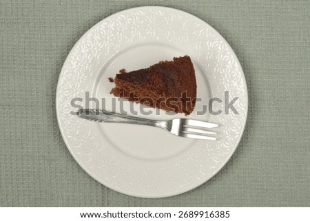 A slice of dark chocolate cake is served on a white patterned plate next to a silver fork on a green textured tablecloth