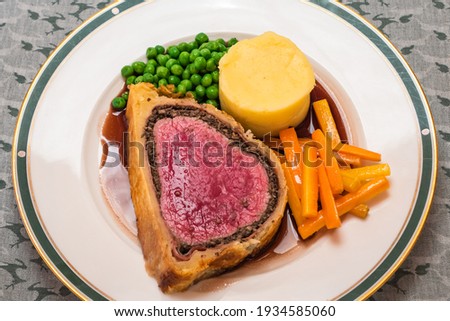 Slice of Fillet of Beef Wellington with Red Wine Gravy and Mashed Potatoes, and Vegetables like Carrots and Peas on a Plate