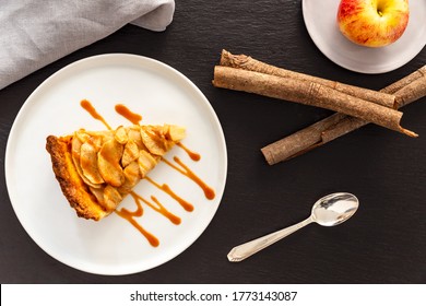 Slice of French apple tart, an open faced apple pie, aside a Gala apple in a small grey plate, a vintage silver spoon, cinnamon sticks and a grey linen napkin on a grey slate background. Flat Lay
