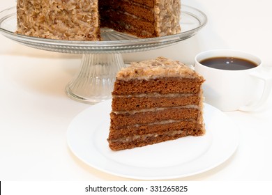 Slice Of German Chocolate Cake Removed From Whole Cake Which Is In Background Along With Cup Of Coffee.  Isolated On White Background.