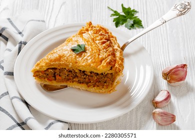 slice of  Ground Beef Meat Pie with a flaky puff pastry double crust with hearty minced beef cooked with vegetables and seasoning in white baking dish on wood table