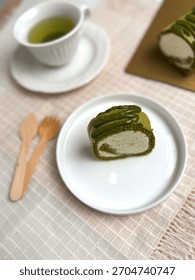 A slice of matcha roll cake served on a white ceramic plate with green tea and wooden cutlery on a beige cloth.Bright,clean tabletop food photography ideal for dessert menus and bakery promotions.