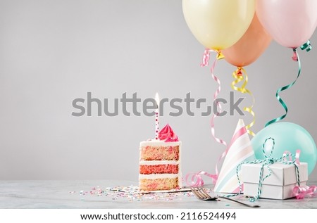 Slice of pink funfetti Birthday cake with candle, presents, hats and colorful balloons over light grey background. Scene from a birthday party!