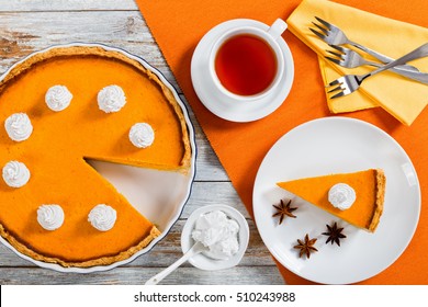 Slice Of Pumpkin Tart - Dessert For Thanksgiving And Christmas, Decorated With Whipped Cream. Cup Of Tea And Whole Pie Cut In Pieces Are On Background, View From Above