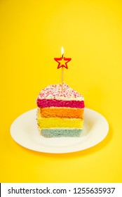 Slice Of Rainbow Cake With Birning Candle In The Shape Of Star On White Round Plate Isolated On Bright Yellow Background. Happy Bithday, Party Concept. Vertical Card. Selective Focus. Copy Space