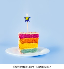 Slice Of Rainbow Cake With Birning Candle In The Shape Of Star On White Round Plate Isolated On Blue Background. Happy Bithday, Party Concept. Square Card. Selective Focus. Copy Space