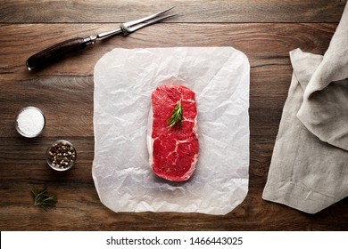 Slice Of Raw New York Steak On White Cooking Paper And Wooden Table. Decorated With Salt, Pepper, Meat Fork And Napkin. Overhead View.