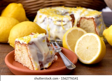 The slice of a sicilian lemon cake with sugar frosting in a ceramic plate, fork, lemons and a straw basket at the background
