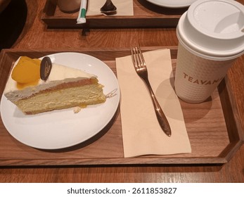 A slice of soft mango cake served on a white plate with a fork and napkin on a wooden tray, accompanied by a warm drink in a cup labeled 