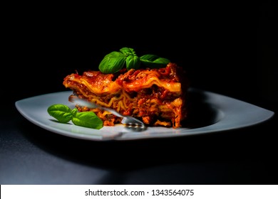 Slice Of Traditional Italian Lasagne With Vegetables, Minced Meat And Cheese And Basil Decoration. On A White Dish And Black Background. The Fork Inserted Into Lasagna