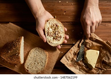 
A slice of wheat bread and butter, sprinkled with various grains and a stained knife in the man’s hands. Homemade butter and sliced ​​fresh bread on a rustic table. Wooden background.