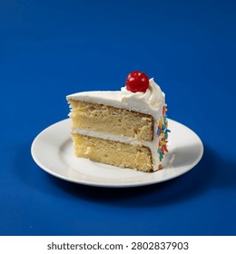 A slice of yellow cake with two layers of white frosting, topped with a cherry and rainbow sprinkles on the side, sits on a white plate against a solid blue background.