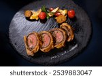 Sliced beef Wellington with glaze and herbs, colorful roasted vegetables on a dark slate plate.