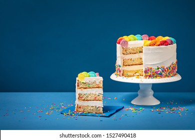 Sliced Confetti Birthday Cake  With Rainbow Colored Icing And Sprinkles Over A Blue Background.