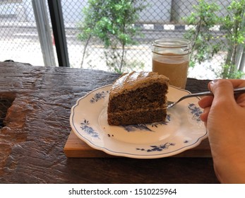 A Sliced Of Earl Grey Cake Served On A Small Plate. A Glass Of Latte. A Hand Is Holding A Fork.