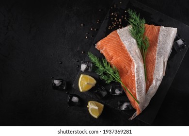sliced fresh raw salmon lying on a stone cutting board with lemon wedges, green dill, spices and ice cubes on a black textured concrete background. top view. beautiful sea food concept with copy space