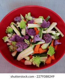 Sliced green spiral Romanesco broccoli, purple cauliflower, orange carrot and zucchini in red bowl