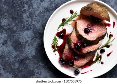 Sliced grilled beef with blueberry sauce on white plate over blue stone background with free space. Tasty medium rare roast beef with berry sauce. Top view, flat lay