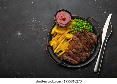 Sliced Grilled Meat Beef Steak With Potato Chips French Fries And Green Peas Served With Red Tomato Sauce On Rustic Metal Plate On Black Stone Background Table From Above With Space For Text