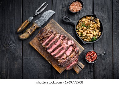 Sliced grilled rib eye beef steak beef marbled rare set, on wooden serving board, with white beans and rosemary in cast iron pan, with meat knife and fork, on black wooden table background, top view 