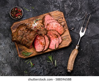 Sliced grilled roast beef with fork for meat on wooden cutting board. Black background. Top view.