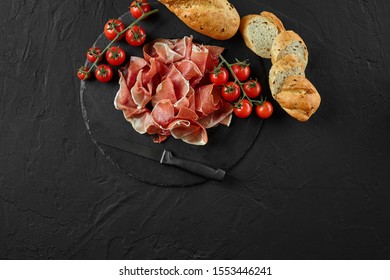 Sliced jamon, cherry tomatoes, sliced baguette and a knife on black stone slate board against a dark grey background. Close-up. Top view. Copy space.