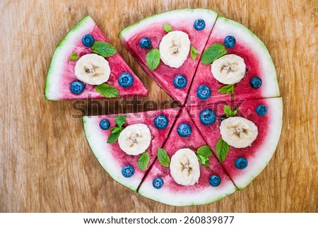Sliced juicy watermelon pizza on wood, closeup view from above. Ingredients are watermelon, blueberries, banana, mint, and coconut shavings.