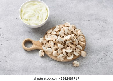 Sliced mini champignon mushrooms on a wooden board and a bowl of chopped onions on a gray textured background. Cooking delicious vegan food.