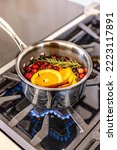 Sliced oranges, cranberries, rosemary and cinnamon sticks simmering on a gas stove with a blue flame in a metal saucepan
