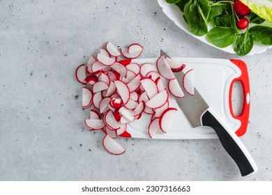 Sliced radishes on a white plastic cutting board, baby spinach, and lettuce on a light gray stone background, close-up view - Powered by Shutterstock