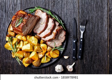 Sliced roasted pork loin served with baked potato wedges, rosemary  on a plate with garlic steak cutlery on a dark wooden background, top view, close up