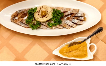 Sliced salted fish herring decorated with a sprig of parsley, onion rings and green olive on an oval white ceramic plate and a plate with mustard and spoon close-up on the tablecloth background - Powered by Shutterstock