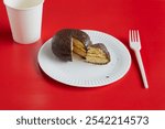 A sliced small piece of chocolate pie from a whole on a white disposable plate with a white paper cup nearby against a red background, a portion of dessert, diet, small serving.