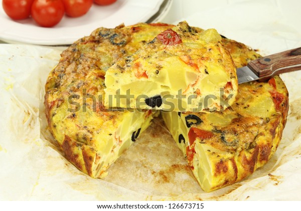 Sliced Spanish potato tortilla with\
black olives and tomatoes on a baking paper with a\
knife
