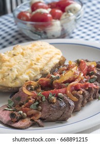 Sliced Steak Covered In A Sauce Of Tomatoes, Peppers, Onions And Seasoning, Accompanied With Twice Baked Potato, And A Bowl Of Caprese Salad In The Background