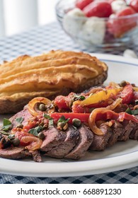 Sliced Steak Covered In A Sauce Of Tomatoes, Peppers, Onions And Seasoning, Accompanied With Twice Baked Potato, And A Bowl Of Caprese Salad In The Background