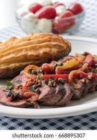 Sliced Steak Covered In A Sauce Of Tomatoes, Peppers, Onions And Seasoning, Accompanied With Twice Baked Potato, And A Bowl Of Caprese Salad In The Background