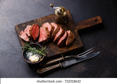 Sliced tenderloin Steak Roast beef with knife and fork carving set on wooden cutting board on dark background