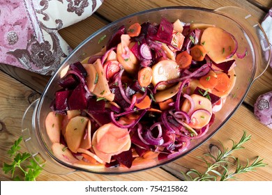 Sliced vegetables (potatoes, red beets, carrots, red onions) in a glass roasting pan on a table, top view