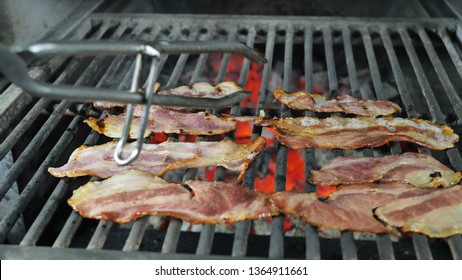 Slices of bacon are grilled, the fat from the bacon drains from the fried bacon, the chef in black gloves with iron tongs picks up the finished bacon from the grill