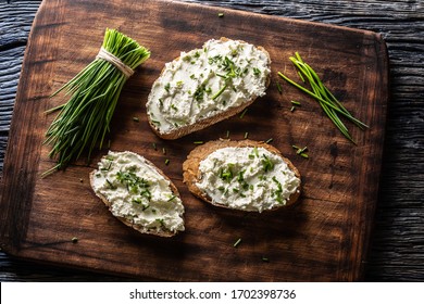 Slices Of Bread With A Cottage Cheese Spread, Freshly Cut Chives, And A Bunch Of Chive Aside Placed On A Dark Brown Cutting Board.