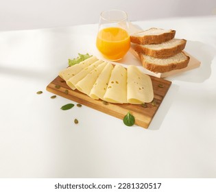 slices of cheese with greens on a wooden board and orange juice with bread behind. maasdam on a light background. yellow cheese under sunlight. dairy products on the table - Powered by Shutterstock