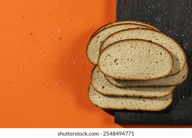 Slices of fresh bread arranged on a dark wooden board with wheat stalks and crumbs scattered against a vibrant orange background. A rustic display perfect for bakery or food-themed concepts - Powered by Shutterstock