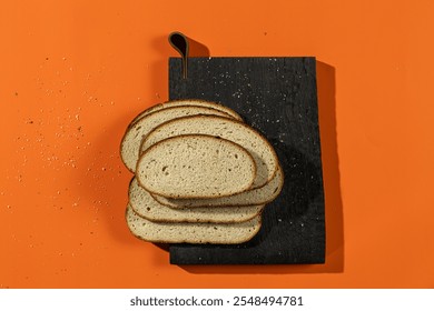 Slices of fresh bread arranged on a dark wooden board with wheat stalks and crumbs scattered against a vibrant orange background. A rustic display perfect for bakery or food-themed concepts - Powered by Shutterstock