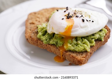 Slices of gluten-free sunflower seeds bread with mashed avocado, poached egg with egg yolk dripping and sesame seeds on white plate on green checkered napkin