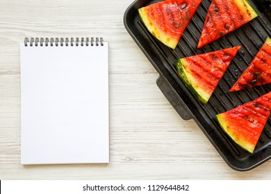 Slices Of Grilled Watermelon In Grilling Pan With Notepad On A White Wooden Surface, From Above. Flat Lay, Overhead, Top View. 