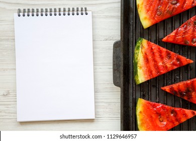 Slices Of Grilled Watermelon In Grilling Pan With Notebook On A White Wooden Background, Top View. Flat Lay, Overhead. 