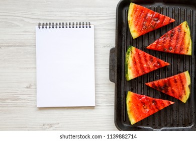 Slices Of Grilled Watermelon In Grilling Pan With Blank Notepad On A White Wooden Surface, Top View. Flat Lay, Overhead. Space For Text.